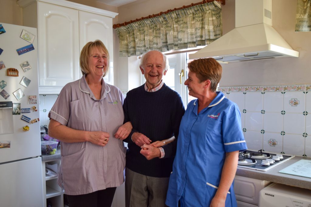 A male resident linking arms with two carers in the kitchen.